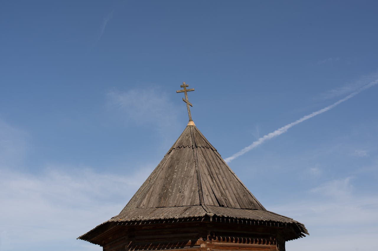 A wooden church roof topped with a cross against a clear blue sky, representing faith.